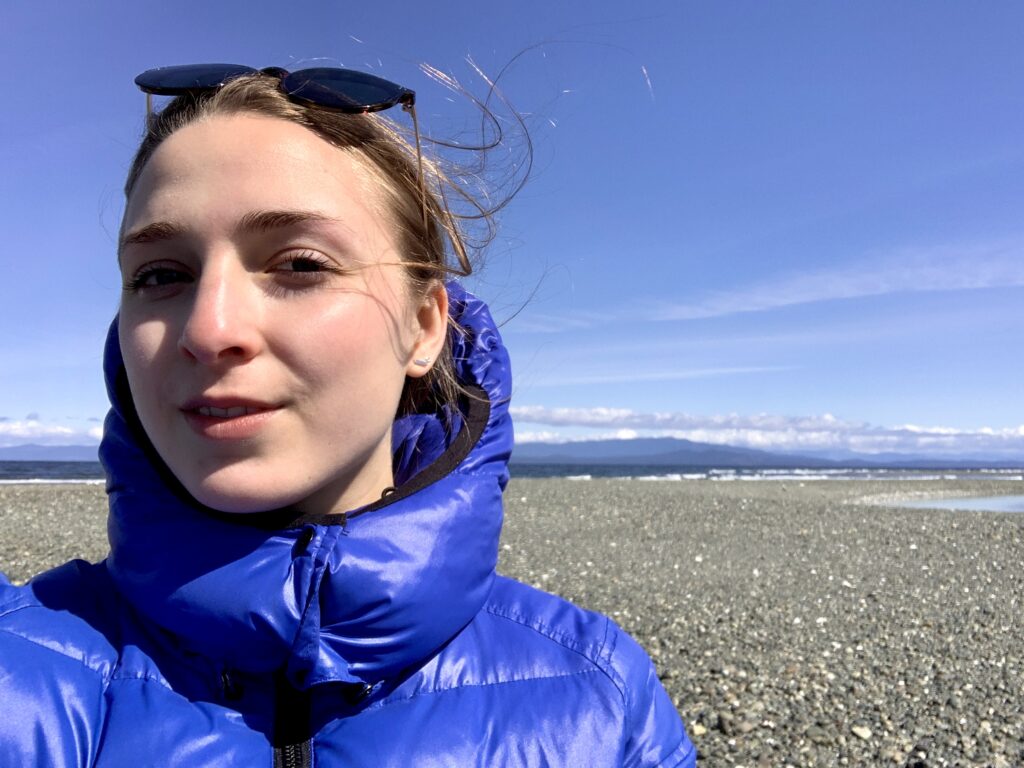A smiling woman in a blue jacket stands on a cold beach. Jenna of Active Travel Life writes about why she started a travel blog.